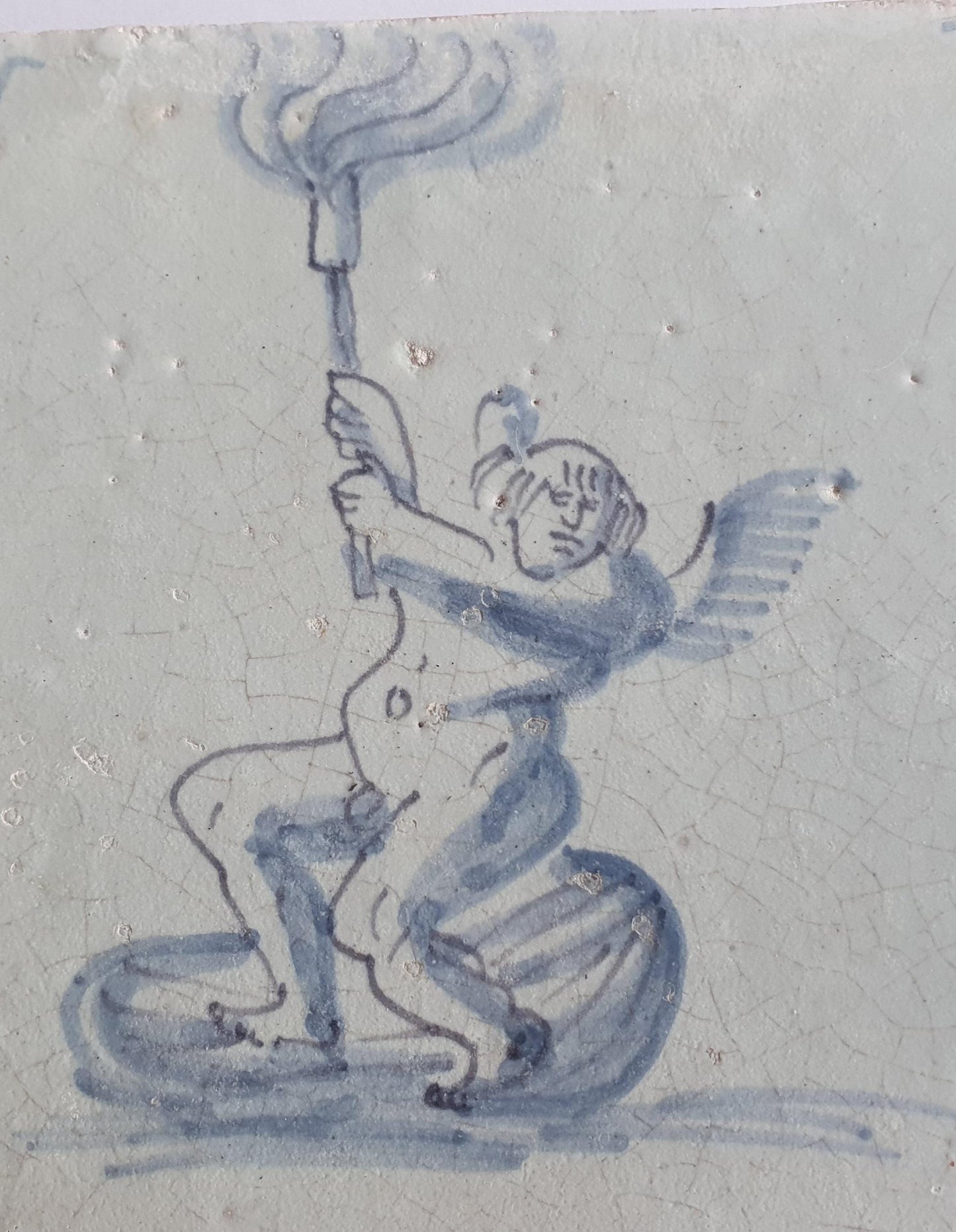 This antique Delft tile depicts Cupid lifting a burning torch in the sky. 17th century