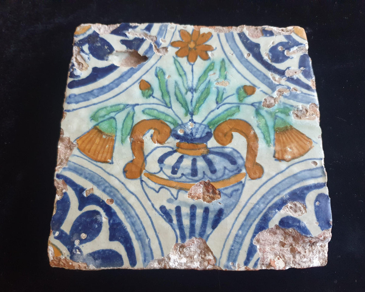 Delft tile from circa 1620 is decorated in polychrome and features a stylized vase containing various stylized flowers. Planttile